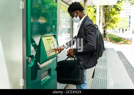 Vista laterale di uomo d'affari afroamericano in maschera protettiva con Valigia usando il terminale ATM di strada nella città di Barcellona Foto Stock