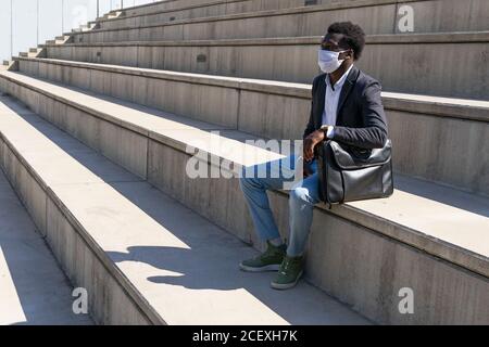 Imprenditore africano americano maschio indossando maschera medica rilassante sulla pietra scale in città durante il coronavirus e guardando lontano Foto Stock