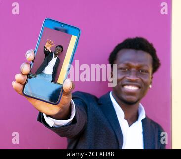 Allegro giovane afroamericano ben vestito imprenditore dimostrare telefono cellulare con autoritratto in piedi contro parete rosa Foto Stock