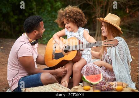 Carina ragazza dai capelli ricci che si diverte e suona la chitarra durante il picnic con i genitori multirazziali nella giornata estiva in natura Foto Stock