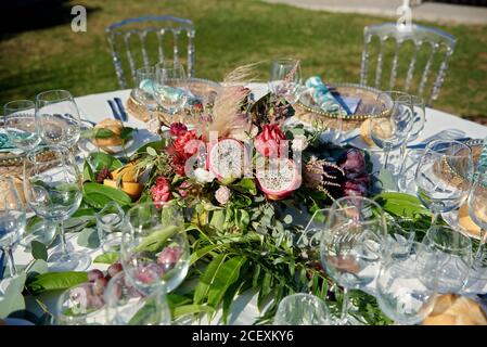 Tavolo rotondo con vari frutti e fiori circondati da sedie e posto in giardino per la celebrazione del matrimonio Foto Stock
