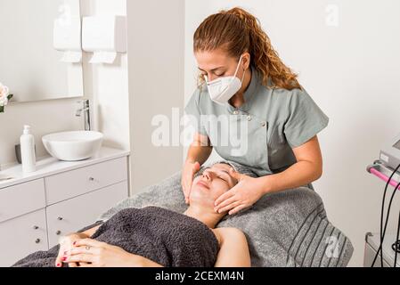 Medico estetista professionista in maschera protettiva applicare crema facciale su Donna mentre si prepara per la procedura dermatologica in moderno salone Foto Stock