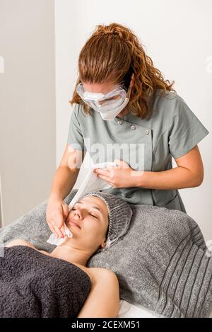 Irriconoscibile specialista femminile in maschera medica e occhiali di protezione che strofinano la faccia di rilassato Donna con asciugamano durante il trattamento di bellezza in clinica moderna Foto Stock