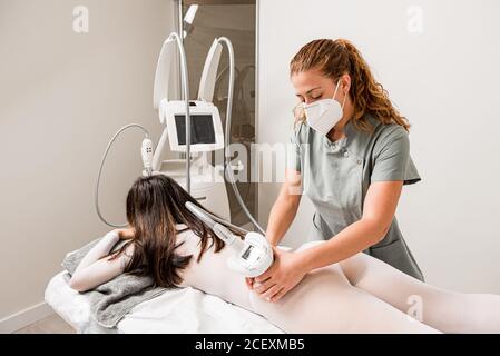 Massaggiouso in maschera protettiva facendo massaggio anti cellulite lipo a. natiche client femminili vestite con tuta bianca durante la cura del corpo procedura in salone di cosmetologia dell'apparato Foto Stock