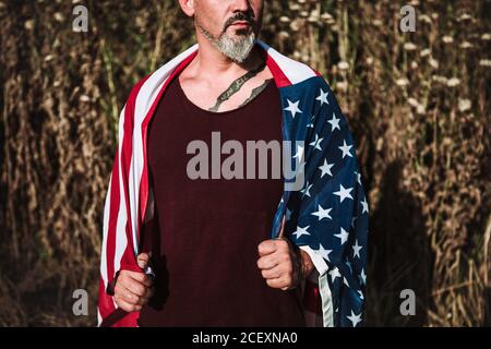 Grave taglio irriconoscibile maschio di media età con tatuaggio avvolto In bandiera americana guardando via mentre si levano in piedi contro la natura offuscata Foto Stock