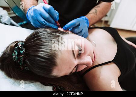 Crop tatuista maschio irriconoscibile che fa segni su orecchio di femmina cliente prima di applicare il tatuaggio durante il lavoro in un salone moderno Foto Stock