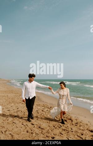 Sposo e sposo giovane e felice a tutta lunghezza in boho elegante abiti che tengono le mani e passeggiano lungo la spiaggia sabbiosa con ondate mare Foto Stock