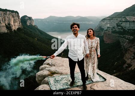 Corpo pieno di felice giovane coppia sposata in elegante abiti che tengono bombe colorate di fumo che tengono le mani mentre celebrano il matrimonio In cima alla roccia a Morro de labella in Spagna Foto Stock