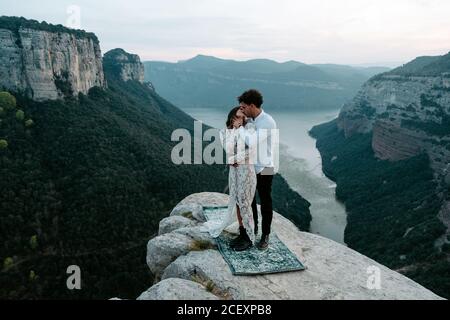 Vista laterale del giovane e della donna in giovane età in abiti eleganti che si ergono in cima alla scogliera rocciosa di Morro de Labella abbracciandosi e baciandosi dolcemente Foto Stock