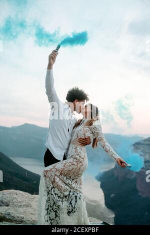 Vista laterale del corpo di una giovane e felice coppia sposata in abiti eleganti che tengono bombe di fumo colorate e baciare mentre Celebrare il matrimonio in cima alla roccia a Morro de labella In Spagna Foto Stock