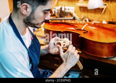 Vista laterale di abile giovane artigiano con intaglio a coltello ponte di violino in legno durante la creazione di strumenti a corda in officina Foto Stock