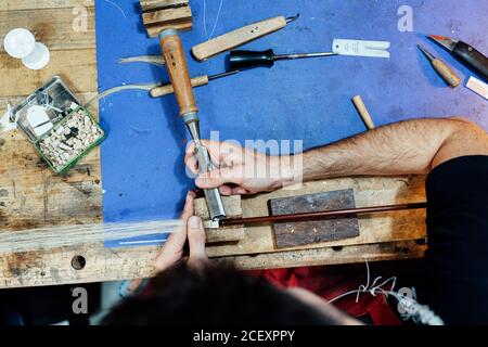 Vista dall'alto del luzier artigianale esperto di raccolto che usa scalpello mentre fare fiocco di violino in officina Foto Stock
