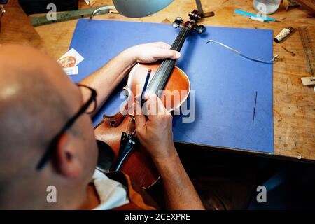 Vista posteriore ad alto angolo di un artigiano che regola le corde ponte di violino mentre si lavora alla scrivania di legno in officina Foto Stock