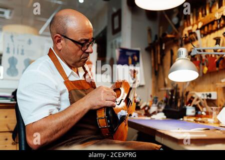 Vista laterale del liutaio maschio maturo in grembiule e bicchieri seduto sulla sedia e tenendo in mano il violino restaurato mentre si lavora officina Foto Stock