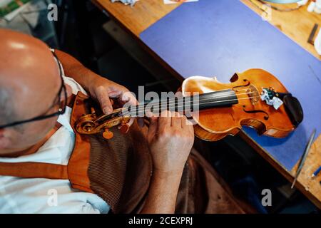 Da sopra irriconoscibile luzier maschio maturo in grembiule e bicchieri seduto sulla sedia e tenendo in mano il violino restaurato mentre si lavora officina Foto Stock