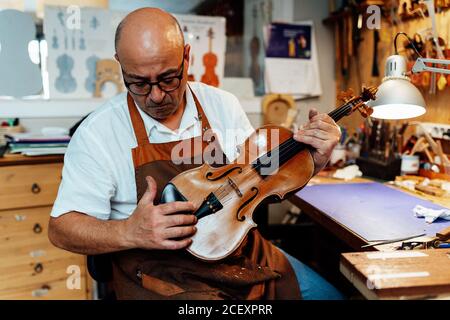 Liutaio maschile maturo in grembiule e bicchieri seduti sulla sedia e tenendo in mano il violino restaurato mentre si lavora in officina Foto Stock