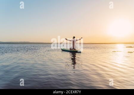 Vista posteriore del corpo intero di uomo irriconoscibile che fa la variazione di yoga asana con braccia aperte mentre galleggia su tavola a pale dentro calmare l'acqua del lago al tramonto Foto Stock