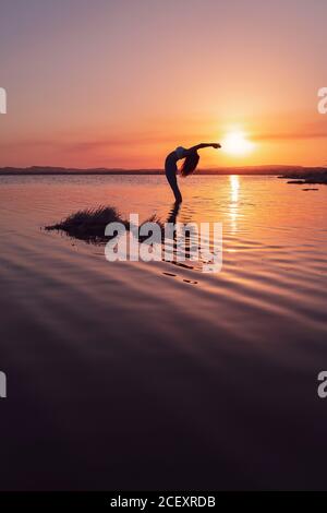 Vista laterale su tutta la lunghezza di una giovane femmina flessibile in abbigliamento sportivo Praticare yoga e fare stare in piedi indietro posa con gli occhi chiusi in acque calme del lago durante il tramonto Foto Stock