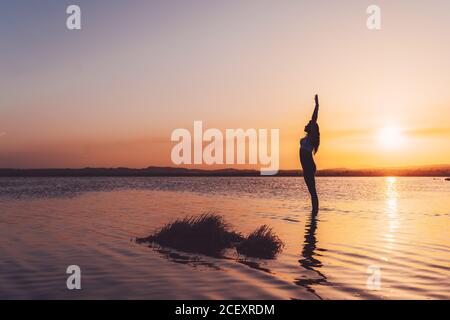 Vista laterale del corpo di una femmina sottile e irriconoscibile in abbigliamento sportivo In piedi in verticale Saluta posa con le braccia in su nel lago acqua mentre si pratica yoga al tramonto Foto Stock