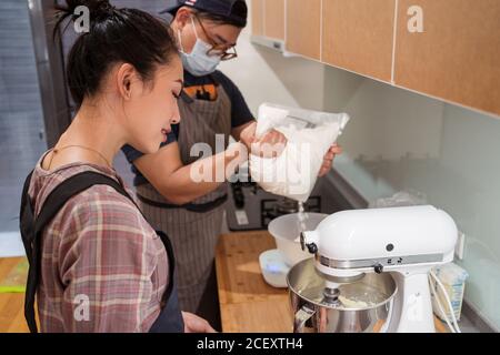Giovane uomo asiatico in maschera pesando zucchero a velo mentre Donna mescolando burro in stand mixer Foto Stock
