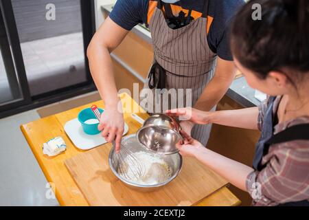 Angolo alto di irriconoscibile Donna e uomo in maschera versando farina in pastella per biscotti sul banco Foto Stock