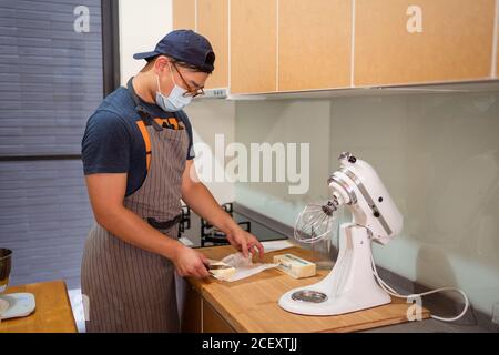 Giovane panettiere maschile in maschera medica e grembiule tagliando burro in pezzi in piedi al banco con miscelatore a cavalletto Foto Stock