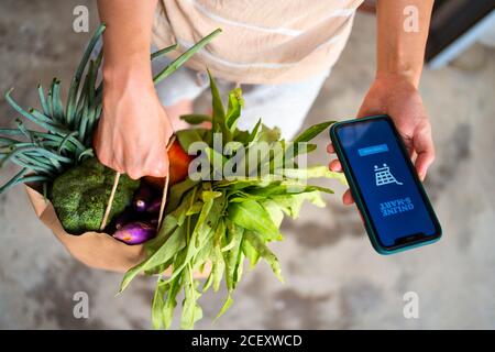 Dall'alto di donna anonima in piedi con sacchetto di carta con frutta e verdura mature consegnate tramite app per lo shopping online Foto Stock