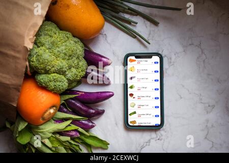 Vista dall'alto di frutta e verdura assortite in sacchetto di carta organizzato sul tavolo con smartphone con app per lo shopping online e consegna Foto Stock