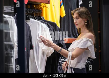 Vista laterale di una giovane donna elegante in piedi vicino al rack e. scegliere la camicia mentre lo shopping in un negozio di abbigliamento moderno Foto Stock