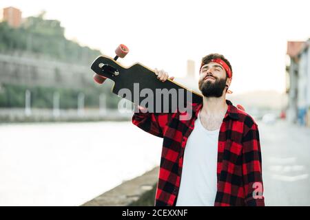 Allegro hipster maschile con cuscinetti in bandana e camicia a scacchi in piedi sul lungomare con longboard e sorridente Foto Stock