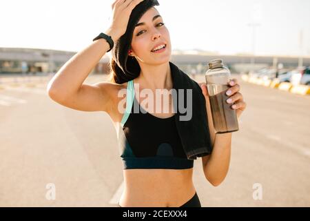 Positivo giovane femmina sottile in sportswear testa di pulizia con asciugamano e bere bevande fresche da bottiglia di plastica mentre si rilassa dopo allenamento duro in strada