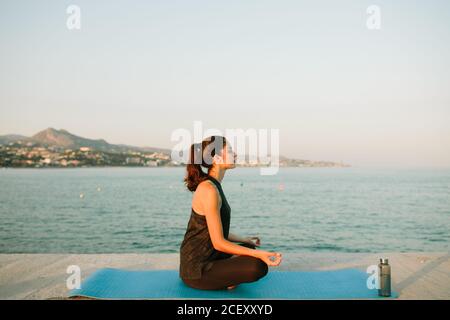 Vista laterale di una donna rilassata seduta sul tappetino yoga Padmasana con mani mudra e la consapevolezza pratica guardando via sfondo di mare al tramonto Foto Stock