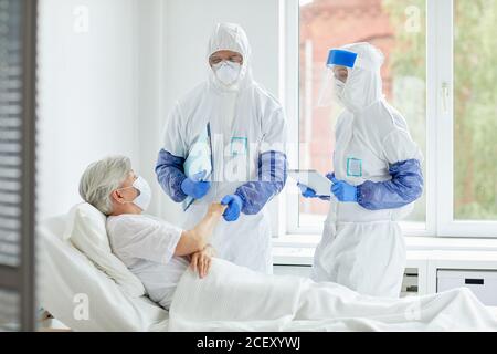 Medici irriconoscibili che indossano tute di protezione con guanti, maschere e occhiali che lavorano con il paziente nel reparto di malattie infettive Foto Stock
