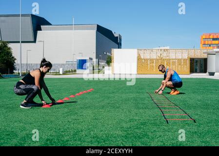 Vista laterale completa del corpo dell'uomo e della donna attivi in abbigliamento sportivo che organizza coni di allenamento e scala di velocità sul prato erboso mentre si prepara per allenarsi insieme allo stadio Foto Stock