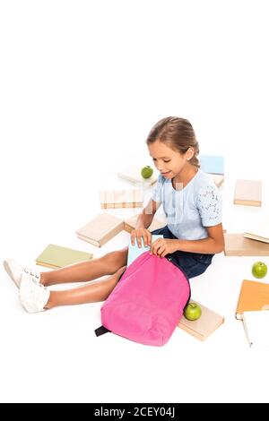 schoolkid seduto vicino alle mele e mettere il libro nello zaino isolato su bianco Foto Stock