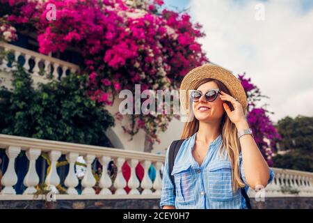 Donna viaggiatore cammina lungo la strada godendo di fiori bougainvillea fioriti. Donna felice che indossa cappello e zaino Foto Stock