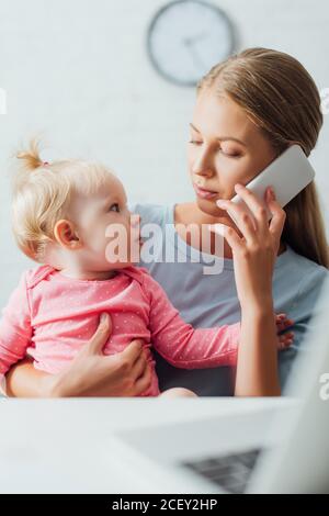 Fuoco selettivo della donna che parla sullo smartphone e che guarda figlia infantile vicino al computer portatile sul tavolo Foto Stock