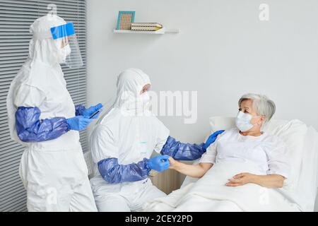 Due medici professionisti che indossano tute protettive con maschere respiratorie e. Guanti esame donna anziana con Covid-19 nel reparto malattie infettive Foto Stock