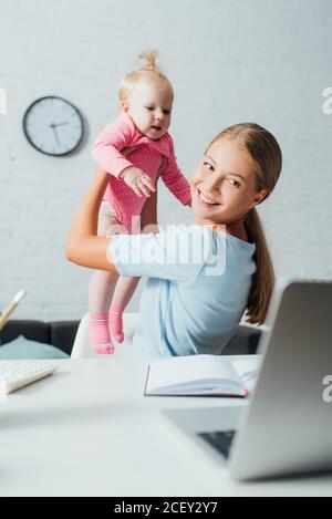 Fuoco selettivo della donna che guarda il laptop mentre tiene il bambino figlia al tavolo Foto Stock