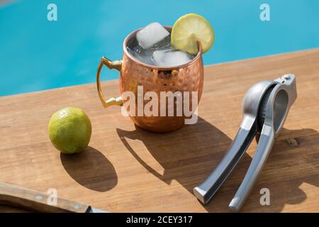 Alto angolo di tradizionale mule halcohol moscovita cocktail con ghiaccio cubetti e lime serviti in una tazza di rame posta con limone spremiagrumi vicino alla piscina Foto Stock