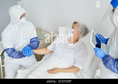 Due operatori medici che indossano tute di protezione con maschere respiratorie e. Guanti che esaminano la salute della donna anziana con Covid-19 in infettivo reparto malattia Foto Stock