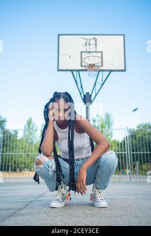 Lunghezza intera di giovane femmina afro-americana con trecce indossate jeans trendy strappati e canotta bianca con sneakers seduto contro la palla da basket sul parco giochi e guardando la macchina fotografica Foto Stock