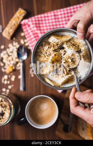 Da sopra di raccolto anonimo persona che tiene ciotola con appetitoso colazione salutare a base di yogurt e fette di banana con farinata d'avena e semi Foto Stock