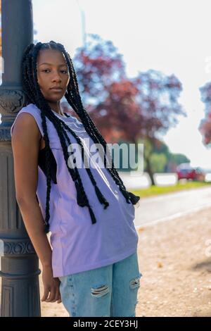 Serious youthful afroamericano femmina con trecce che indossa camicia casual e jeans strappati appoggiati sulla colonna e guardando la fotocamera in parco urbano in estate Foto Stock