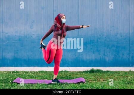 Atleta araba in maschera medica e hijab in piedi mat e allungare le gambe durante il riscaldamento prima dell'allenamento Foto Stock