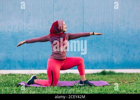 Vista laterale della femmina araba in abbigliamento attivo e hijab Pratica yoga a Crescent Lunge posa sul tappetino nel parco Foto Stock