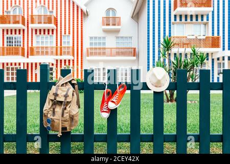 Colorate sneakers rosse del viaggiatore con cappello e zaino appeso Su recinto contro le case a strisce di Costa Nova Foto Stock