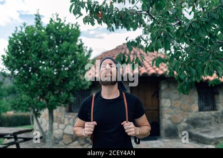 Calma turista maschile con zaino in piedi vicino casa in pietra in insediamento Foto Stock