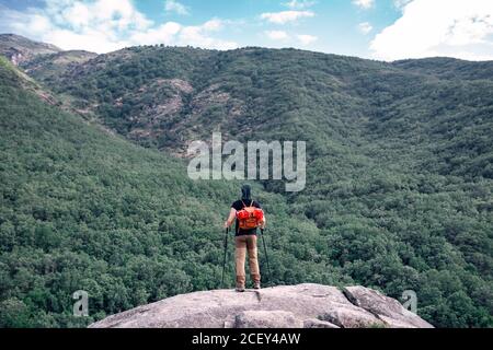 Vista posteriore di un anonimo escursionista maschile con bastoni da trekking in piedi sulla roccia e ammirando la vista di spettacolari alture coperte verde foresta Foto Stock