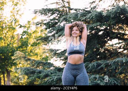 Basso angolo di positivo giovane etnico femminile con Afro capelli con top grigio e leggings che si allungano con le braccia dietro dirigiti accanto a alberi verdi durante l'allenamento nel parco Foto Stock